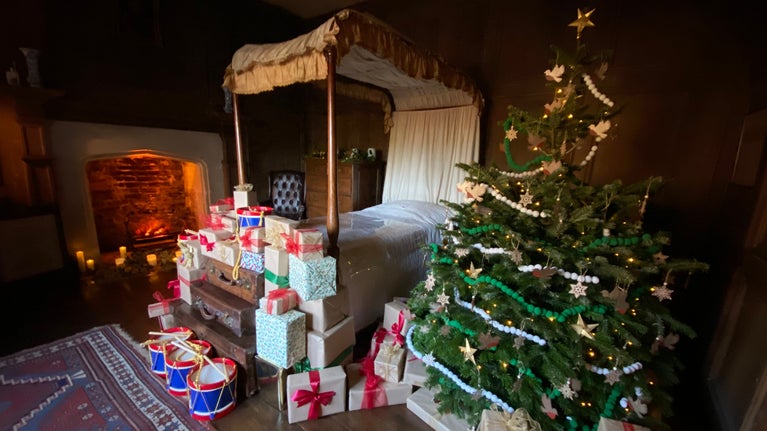 View across a large bedroom with a four poster bed, decorate Christmas tree and gifts under and a lit fire